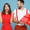 Feliz casal jovem de amigos (homem e mulher) em roupas vermelhas e brancas. Homem segura caixa de presente com laço de fita e mulher faz gesto de vencedora. Isolados sobre fundo azul. Conceito de Dia do Amigo