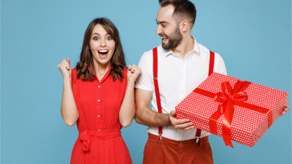 Feliz casal jovem de amigos (homem e mulher) em roupas vermelhas e brancas. Homem segura caixa de presente com laço de fita e mulher faz gesto de vencedora. Isolados sobre fundo azul. Conceito de Dia do Amigo