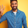 Jovem vestindo camisa jeans, em pé, sobre fundo amarelo isolado, mostrando e apontando para cima com três dedos enquanto sorri confiante e feliz.