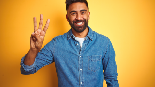 Jovem vestindo camisa jeans, em pé, sobre fundo amarelo isolado, mostrando e apontando para cima com três dedos enquanto sorri confiante e feliz.