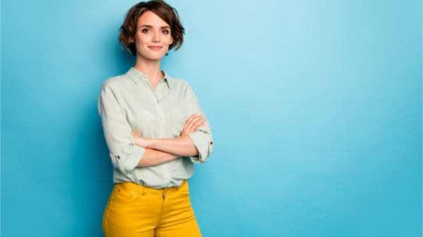 Foto de uma mulher de negócios de cabelo curto, amigável, sorridente, pessoa responsável, braços cruzados, vestindo camisa verde casual e calça amarela, isolada sobre fundo de cor azul.