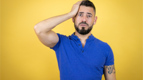 Homem com barba, vestindo camisa polo azul sobre fundo amarelo, colocando uma mão na cabeça e sorrindo como se tivesse esquecido algo.