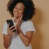 Adorável jovem com smartphone moderno nas mãos, sentindo-se feliz em receber mensagem, olhando para a tela do dispositivo e sorrindo em pé sobre a parede do estúdio bege.