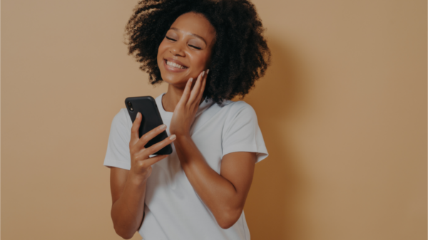 Adorável jovem com smartphone moderno nas mãos, sentindo-se feliz em receber mensagem, olhando para a tela do dispositivo e sorrindo em pé sobre a parede do estúdio bege.