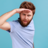 Retrato de homem barbudo adulto jovem bonito olhando longe com a mão sobre a cabeça, vendo algo agradável. Foto de estúdio interior isolada sobre fundo azul.