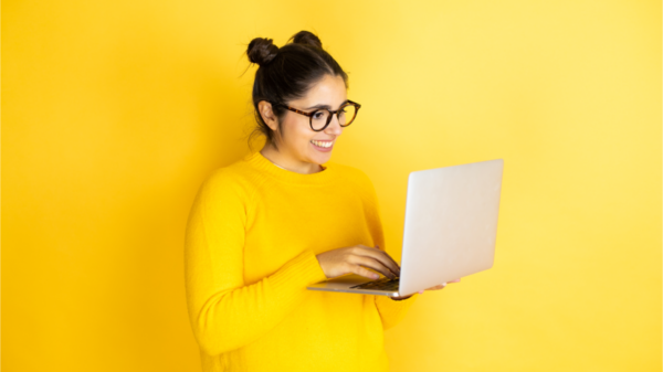 Jovem morena trabalhando usando computador sobre fundo amarelo com um sorriso no rosto, de pé e com um sorriso confiante, mostrando os dentes.