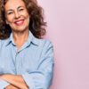 Mulher de meia idade vestindo camisa jeans casual, em pé sobre fundo rosa. Rosto feliz, sorrindo com os braços cruzados, olhando para a câmera. Pessoa positiva.