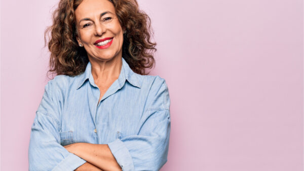 Mulher de meia idade vestindo camisa jeans casual, em pé sobre fundo rosa. Rosto feliz, sorrindo com os braços cruzados, olhando para a câmera. Pessoa positiva.