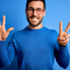 Jovem com barba, vestindo suéter casual e usando óculos sobre fundo azul, mostrando e apontando para cima com os dedos fazendo o número sete enquanto sorri confiante e feliz.