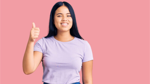 Jovem garota vestindo roupas casuais, sorrindo feliz e positiva, com o polegar para cima, fazendo sinal de aprovação.
