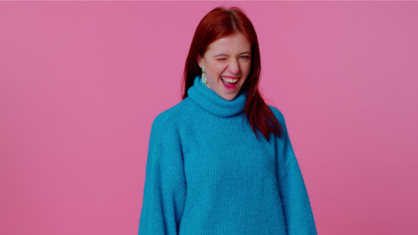 Adolescente ruiva sorrindo e feliz, de suéter azul, piscando o olho, olhando para a câmera dando um sorriso, piscando e flertando, expressando otimismo. Jovem mulher adulta no fundo do estúdio rosa.