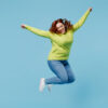 Mulher jovem, alegre e animada vestindo blusa verde, dando um salto alto com as mãos estendidas, isolada no no fundo azul liso. Conceito de estilo de vida de pessoas.
