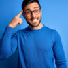 Jovem com barba vestindo suéter casual e usando óculos sobre fundo azul. Sorrindo e apontando para a cabeça com um dedo. Ótima ideia ou pensamento, boa memória, inteligência.