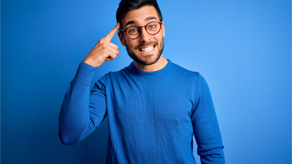 Jovem com barba vestindo suéter casual e usando óculos sobre fundo azul. Sorrindo e apontando para a cabeça com um dedo. Ótima ideia ou pensamento, boa memória, inteligência.