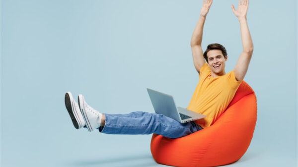 Vista lateral de corpo inteiro de jovem divertido de cerca de 20 anos vestindo camiseta amarela, sentado no pufe, segurando o pc, esticando as mãos e as pernas, isolado no estúdio de fundo azul claro pastel simples.