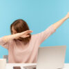 Mulher muito feliz mostrando gesto de dança "dab", realizando meme de sucesso na internet, sentada no local de trabalho com laptop, em escritório em casa. Foto de estúdio interno isolado em fundo azul.
