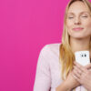Jovem romântica e sonhadora segurando um telefone celular apertado contra o peito com um sorriso de prazer e com os olhos fechados em êxtase, sobre um fundo rosa.