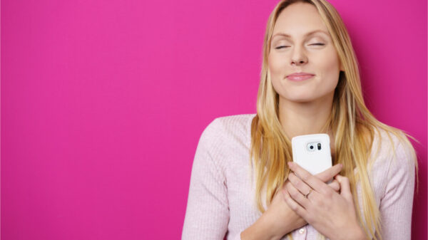 Jovem romântica e sonhadora segurando um telefone celular apertado contra o peito com um sorriso de prazer e com os olhos fechados em êxtase, sobre um fundo rosa.