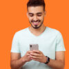 Retrato de homem moreno, alegre, com barba, de camiseta branca, usando celular e sorrindo. Isolado em fundo laranja.