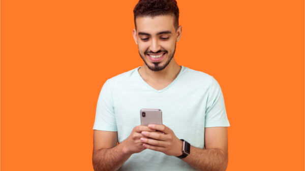 Retrato de homem moreno, alegre, com barba, de camiseta branca, usando celular e sorrindo. Isolado em fundo laranja.