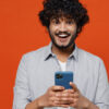 Jovem sonhador, divertido, barbudo, de cerca de 20 anos, vestindo camisa azul, usando celular, isolado em fundo vermelho simples. Retrato de estúdio.