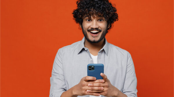 Jovem sonhador, divertido, barbudo, de cerca de 20 anos, vestindo camisa azul, usando celular, isolado em fundo vermelho simples. Retrato de estúdio.