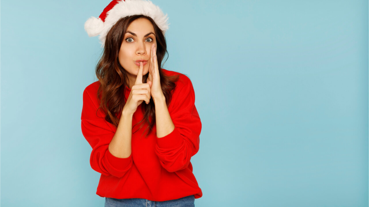 Mulher jovem vestida de Mamãe Noel fazendo gesto de silêncio sobre o espaço de fundo azul. Conceito de segredo de Natal.