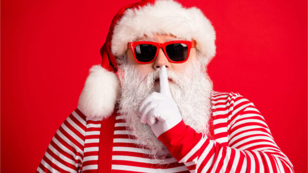Retrato de Papai Noel misterioso de cabelos brancos, usando óculos escuros, fazendo sinal de silêncio, mudo, silencioso, isolado sobre fundo de cor vermelha.