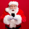 Retrato de homem vestido de Papai Noel usando celular, isolado sobre fundo de cor vermelha.