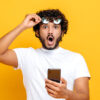 homem em fundo amarelo com um óculos na mão direita e um celular na mão esquerda. Sua cara está pasma