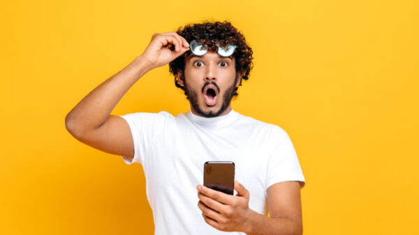 homem em fundo amarelo com um óculos na mão direita e um celular na mão esquerda. Sua cara está pasma