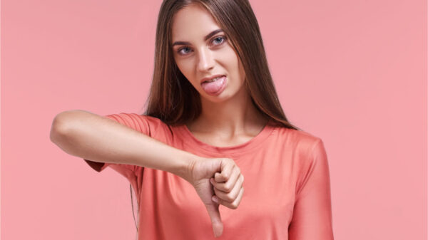 Mulher descontente mostrando sinal de desaprovação, mantendo o polegar para baixo, expressando desagrado, franzindo a testa em descontentamento e mostrando a língua, isolada sobre fundo rosa. Conceito de linguagem corporal.