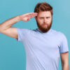 Retrato de homem barbudo responsável saudando com respeito como se fosse um soldado esperando ordem do comandante, obedecendo à disciplina. Foto de estúdio isolado no fundo azul.