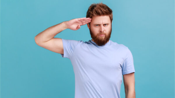 Retrato de homem barbudo responsável saudando com respeito como se fosse um soldado esperando ordem do comandante, obedecendo à disciplina. Foto de estúdio isolado no fundo azul.
