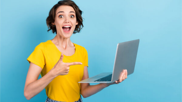 Foto de uma garota segurando netbook com a boca aberta, usando camiseta amarela, isolada no fundo azul.