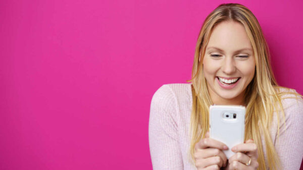 Mulher loira feliz olhando para seu celular com um sorriso surpreso contra um fundo rosa no estúdio.