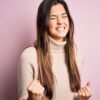 Menina vestindo suéter de gola alta casual, em pé sobre fundo rosa, isolada, muito feliz e animada, fazendo gesto de vencedora com os braços levantados e sorrindo.