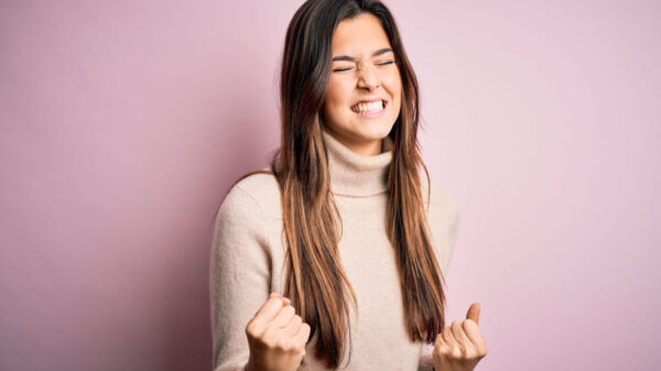 Menina vestindo suéter de gola alta casual, em pé sobre fundo rosa, isolada, muito feliz e animada, fazendo gesto de vencedora com os braços levantados e sorrindo.