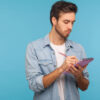 Retrato de homem de camisa jeans fazendo anotações em caderno, escrevendo ideias de negócios, planos futuros, marcando compromissos na agenda. Foto de estúdio interior isolada em fundo azul.