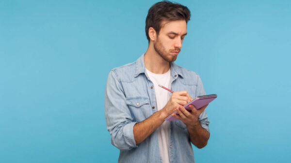 Retrato de homem de camisa jeans fazendo anotações em caderno, escrevendo ideias de negócios, planos futuros, marcando compromissos na agenda. Foto de estúdio interior isolada em fundo azul.
