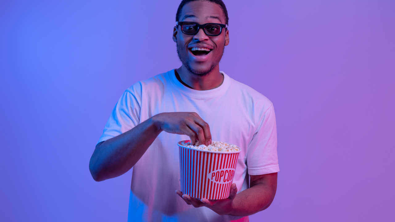 Retrato de homem de óculos 3D com balde de pipoca. Homem alegre olhando para a câmera com entusiasmo, curtindo um bom filme, parado sob a luz neon sobre fundo roxo.