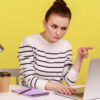 Mulher séria no escritório sentada no local de trabalho, repreendendo algo ou algém, olhando para a tela do laptop. Foto de estúdio interno isolado em fundo amarelo.
