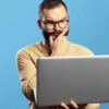 Retrato de um homem barbudo e duvidoso segurando laptop, isolado sobre fundo azul.