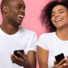 Casal jovem e feliz rindo e usando smartphones isolados no fundo do estúdio rosa. Amigos alegres segurando aparelhos de telefones celulares e se divertindo com celulares juntos.