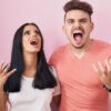 Jovem casal em pé sobre fundo rosa gritando com expressão agressiva e braços levantados. Conceito de frustração.