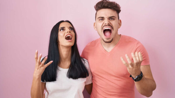 Jovem casal em pé sobre fundo rosa gritando com expressão agressiva e braços levantados. Conceito de frustração.