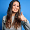 Jovem mulher morena vestindo suéter casual, em pé, sobre fundo azul, feliz, com polegar para cima, fazendo gesto com a mão. Expressão de aprovação olhando para a câmera, mostrando sucesso.