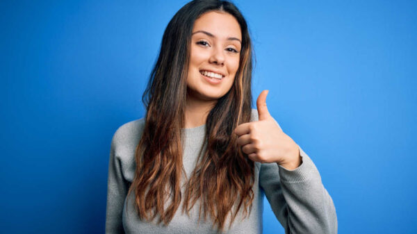 Jovem mulher morena vestindo suéter casual, em pé, sobre fundo azul, feliz, com polegar para cima, fazendo gesto com a mão. Expressão de aprovação olhando para a câmera, mostrando sucesso.