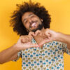 Jovem em pé sobre fundo amarelo, sorrindo, fazendo forma de símbolo de coração com as mãos. Conceito romântico.
