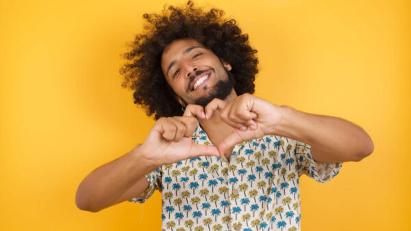 Jovem em pé sobre fundo amarelo, sorrindo, fazendo forma de símbolo de coração com as mãos. Conceito romântico.